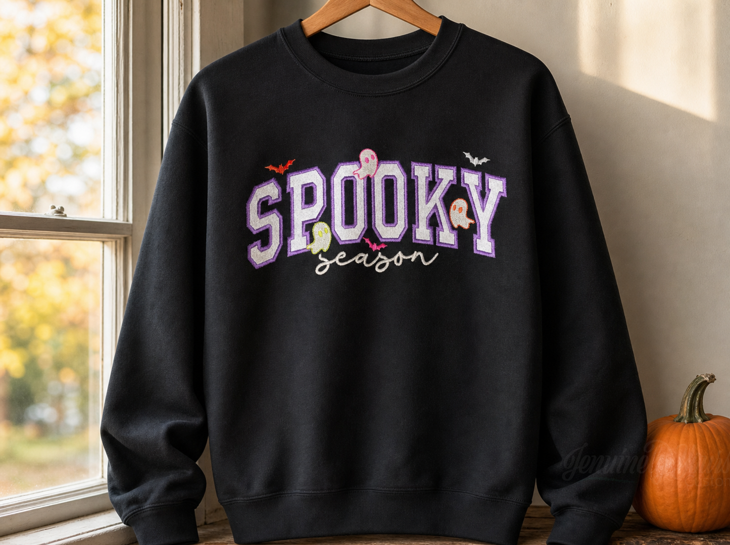 A black crewneck sweatshirt hanging on a wooden hanger, featuring a 'SPOOKY season' embroidered outlined letters and small colorful cartoon ghosts and bats. The sweatshirt is styled on a windowsill next to a small orange pumpkin.