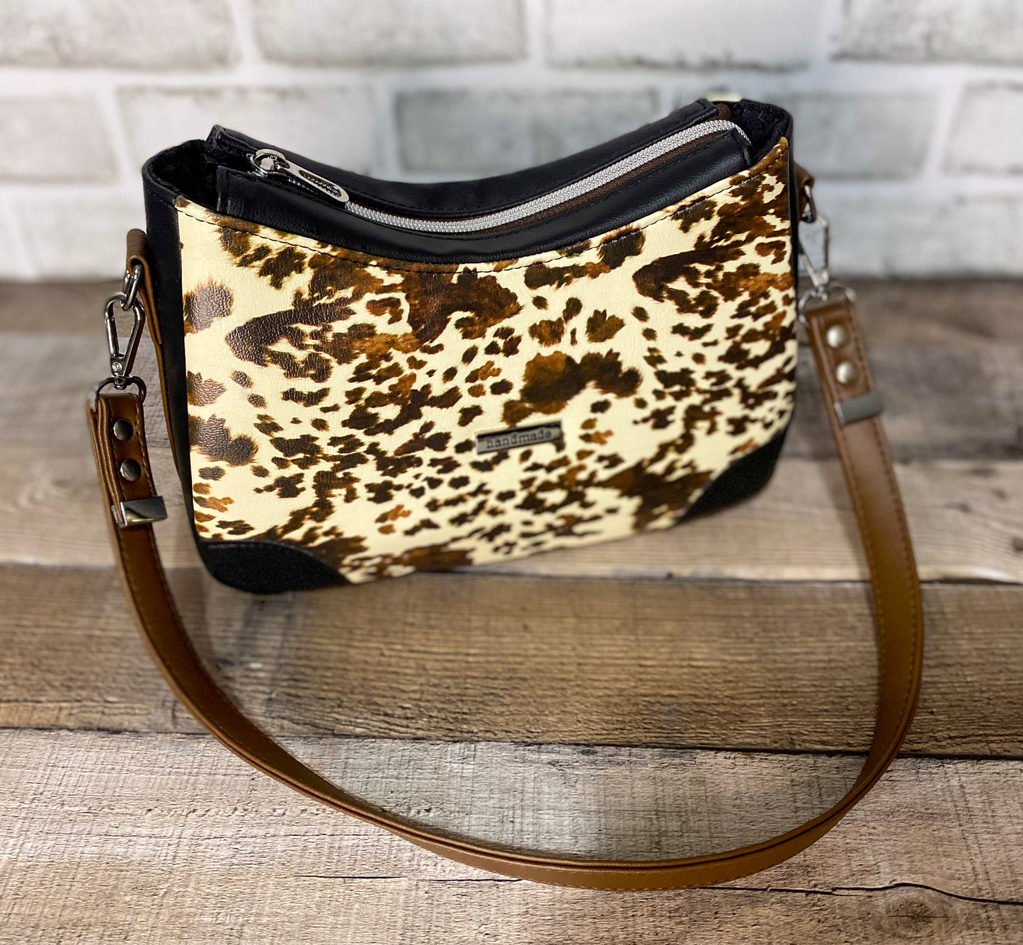 Brown cow print handbag with black faux leather accents. Front view showing the top of the zipper panel. 