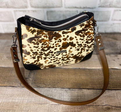 Brown cow print handbag with black faux leather accents. Front view showing the top of the zipper panel. 