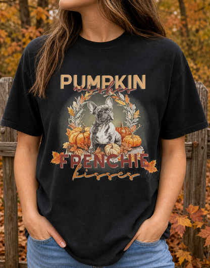 A woman wearing a black graphic t-shirt with a French Bulldog and pumpkin design, standing outdoors in front of a wooden fence with autumn trees.