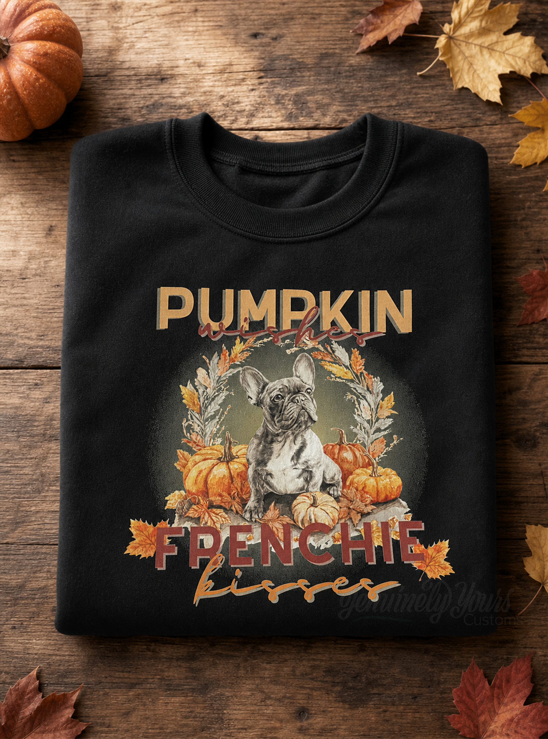 A folded black t-shirt with a 'Pumpkin and Frenchie DTF graphic, displayed on a rustic wooden background with autumn leaves.