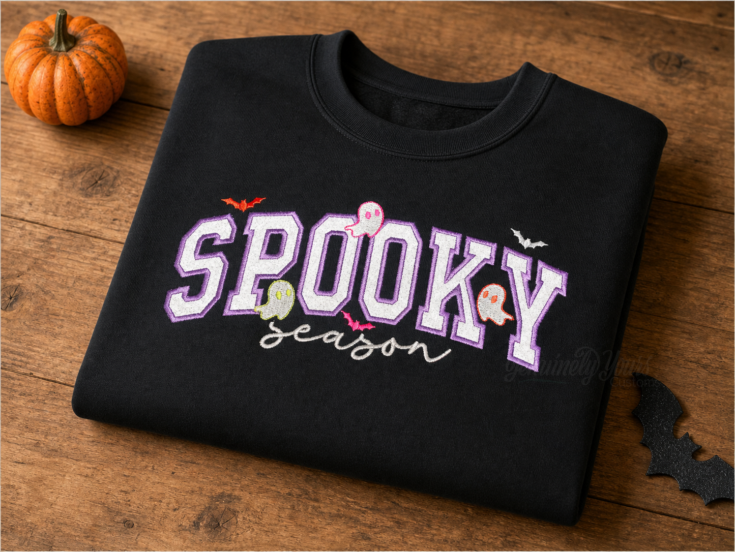 A black crewneck sweatshirt with a 'Spooky Season' emroidered design, folded on a wooden surface next to a small orange pumpkin and a black bat silhouette.