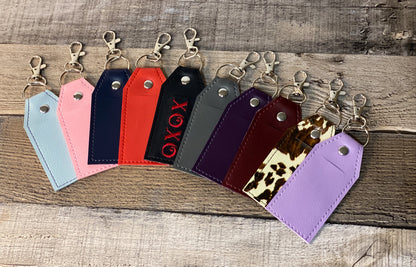 Collection of colorful faux leather chapstick holder keychains on a wooden surface with a brick wall background.