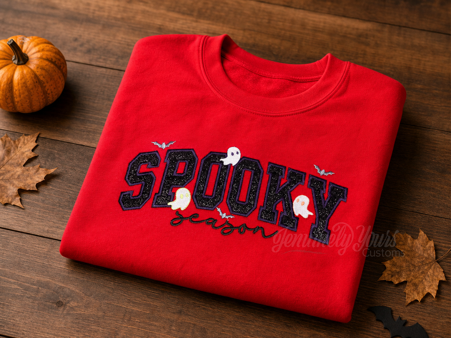 A folded red crewneck sweatshirt with a 'Spooky Season' embroidered in purple with black glitter htv lettering, decorated with small white and orange ghosts and bats, displayed on a wooden table with a small pumpkin and autumn leaves.