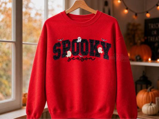 A red crewneck sweatshirt hanging on a wooden hanger, featuring a black 'SPOOKY' embroidered text design with white and orange ghost details and the word 'season' in script below, set against a cozy autumn-themed indoor background.