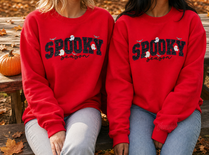 Two women sitting on a wooden bench outdoors wearing matching red crewneck sweatshirts with 'SPOOKY season' embroidered in purple and across the chest.