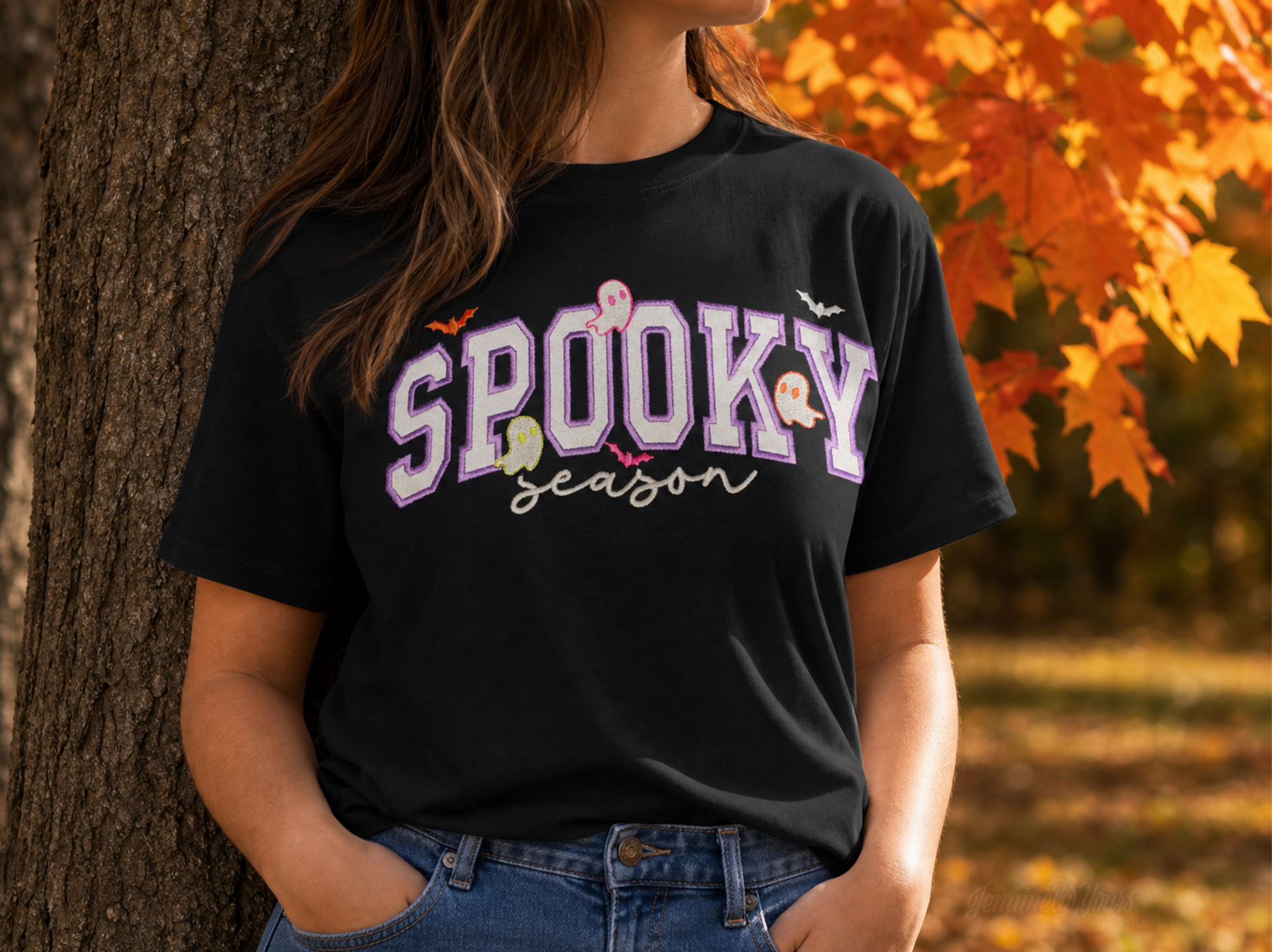 A woman wearing a black graphic t-shirt with 'Spooky Season' embroidered  in purple and white glitter htv letters, posed outdoors next to a tree with autumn leaves in the background.