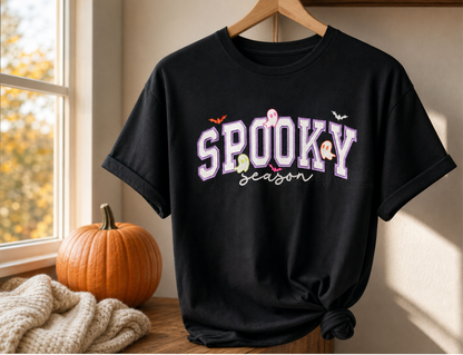 A black short-sleeve T-shirt hanging on a wooden hanger in a warm indoor setting. The shirt has a large 'SPOOKY season' embroidered in the front with purple stitching with white glitter appliqué letters, small orange and white ghost details, and tiny bat silhouettes.