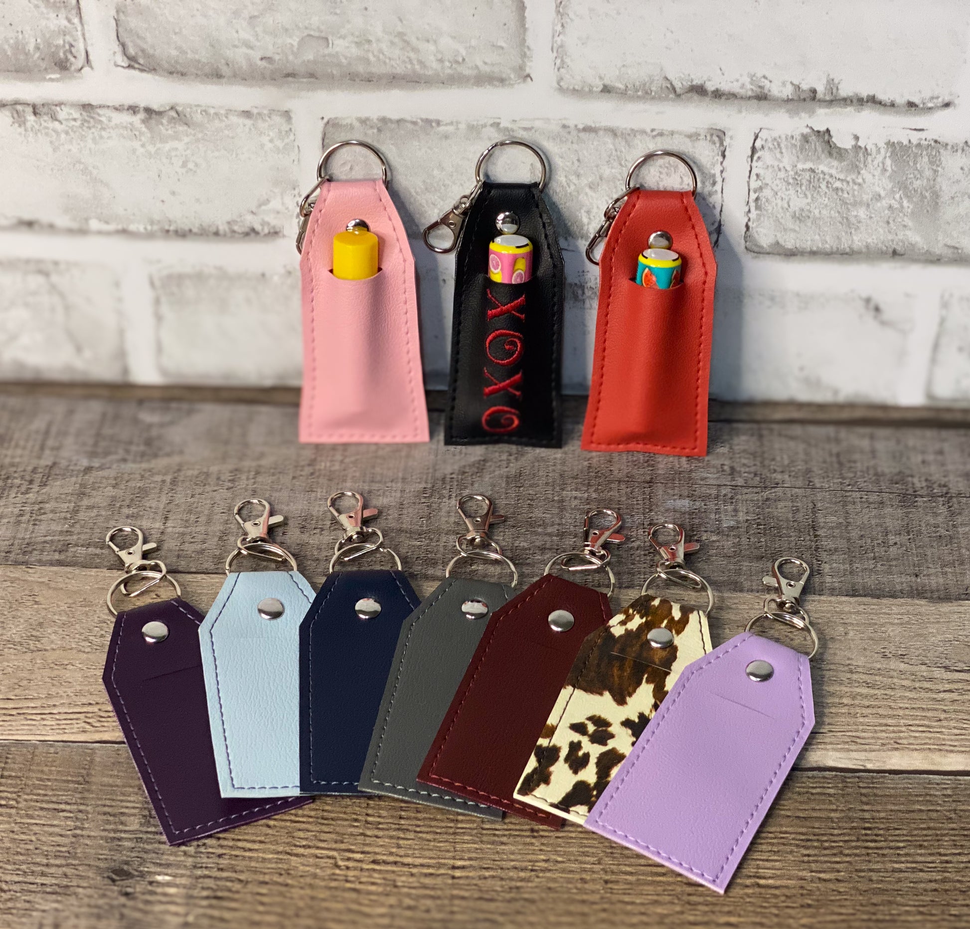 Collection of colorful faux leather chapstick holder keychains on a wooden surface with a brick wall background.