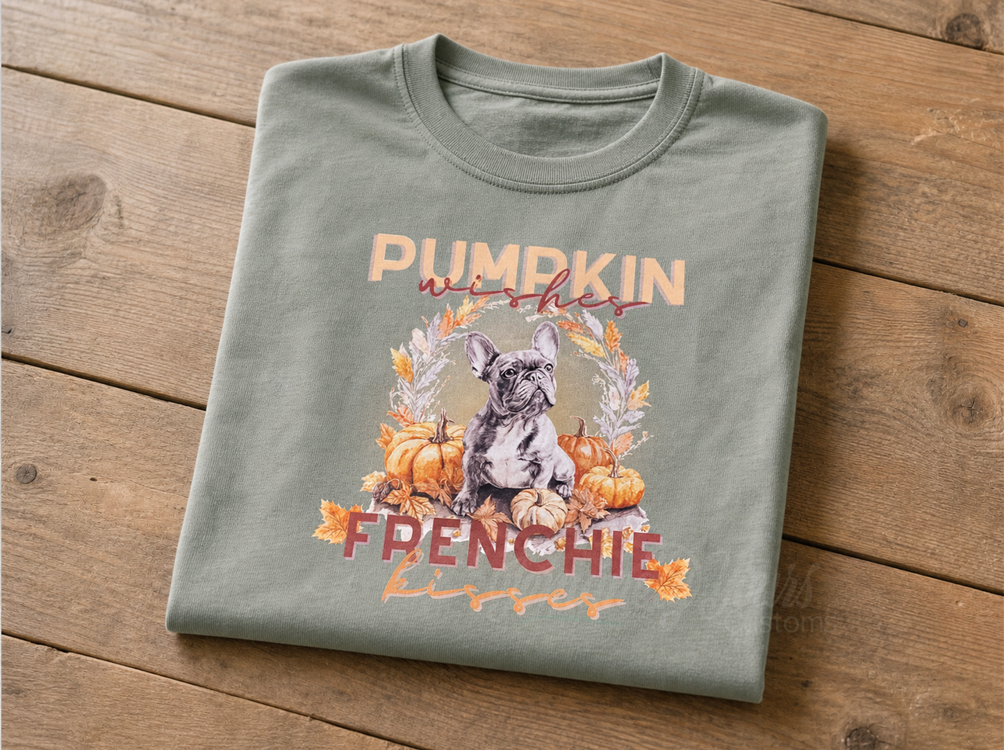 A folded sage green t-shirt with a graphic featuring a French Bulldog, displayed on a rustic wooden background.